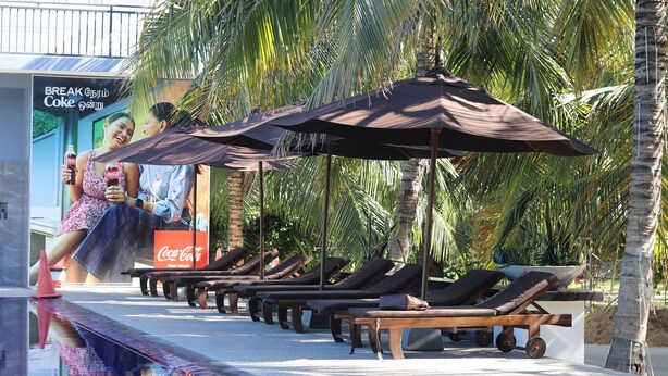 A poolside area with lounge chairs and umbrellas, a Coca-Cola advertisement, and palm trees in the background. - ReeCha Mullai Resort (Iyakachchi)