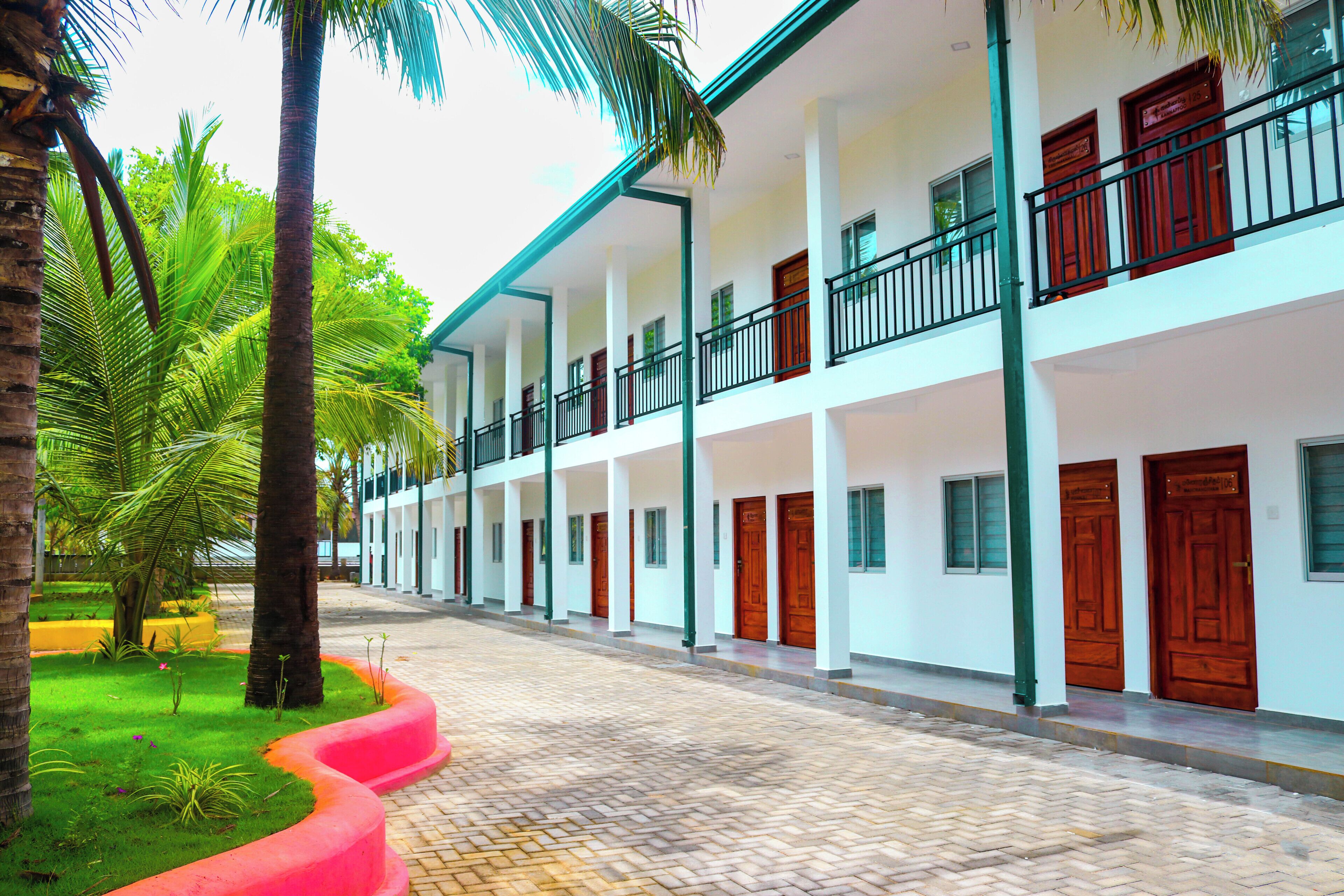 A multi-story building with a central courtyard, featuring a brick pathway, palm trees, and a red flower bed.