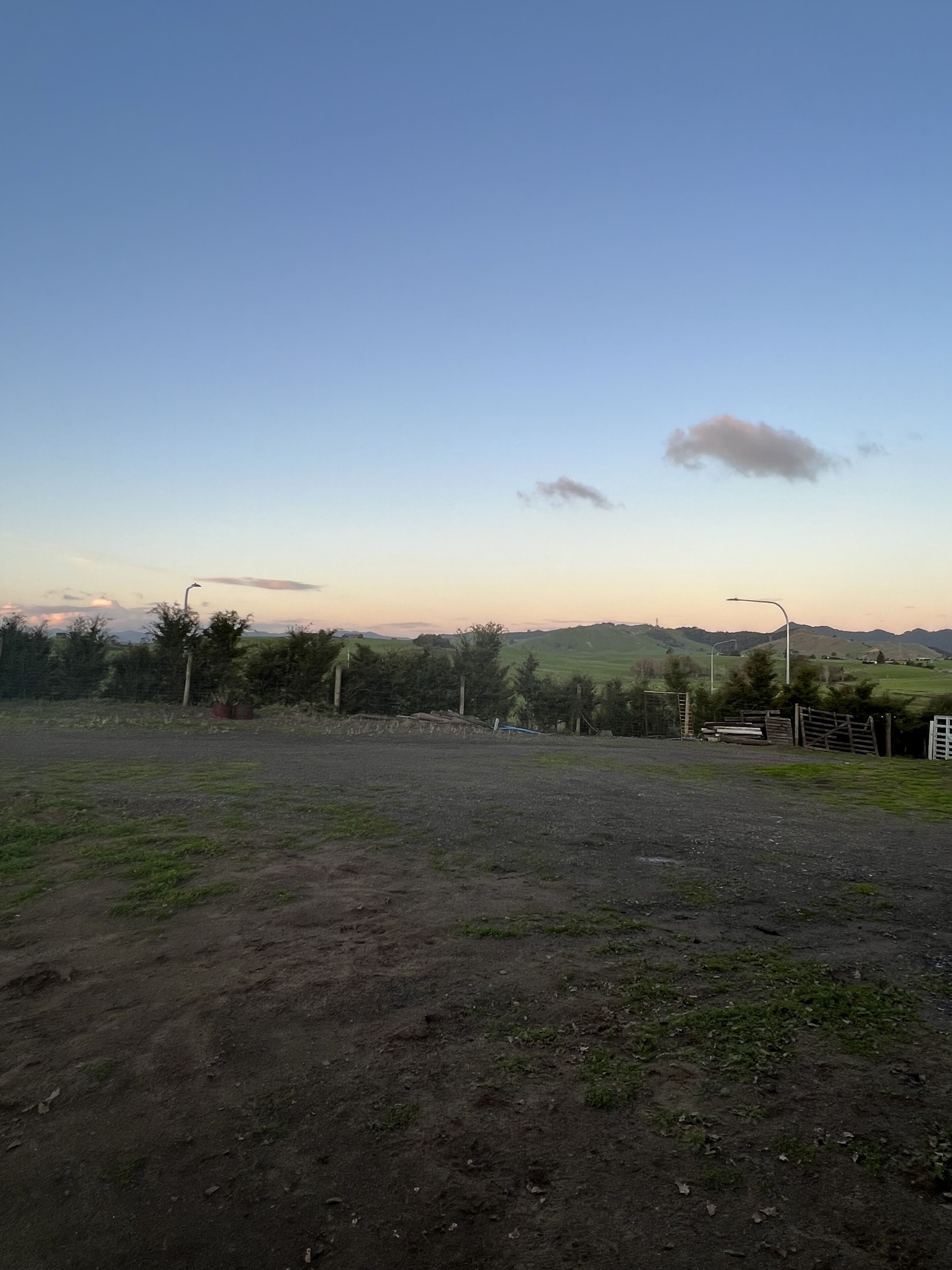Ländliche Landschaft mit einem Feld, spärlicher Vegetation und weit entfernten Hügeln unter blauem Himmel.
