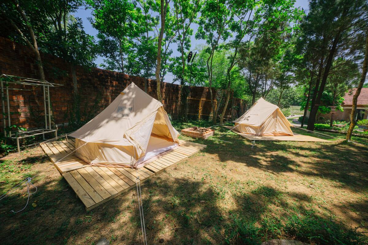 Two beige tents set up on wooden platforms in a wooded area with a brick wall in the background.