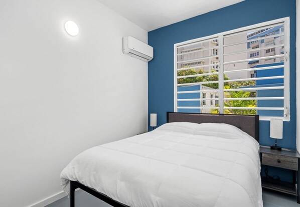 Une chambre à coucher comprenant un lit, un appareil de climatisation, une fenêtre avec des stores et une vue sur les bâtiments et la verdure environnante.