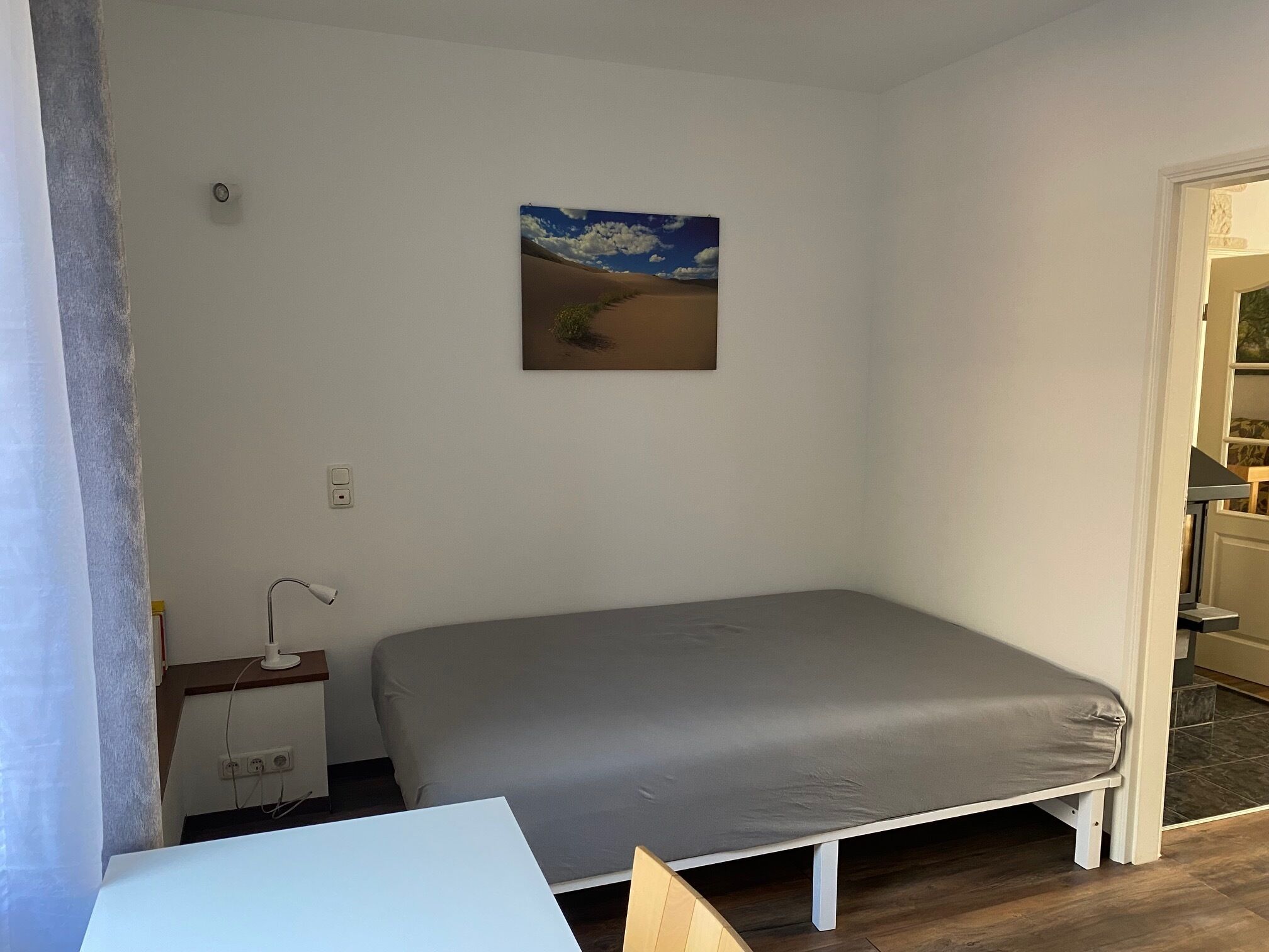 A small, single-bed room with a simple bed, a bedside table, a lamp, and a painting of a desert landscape on the wall.