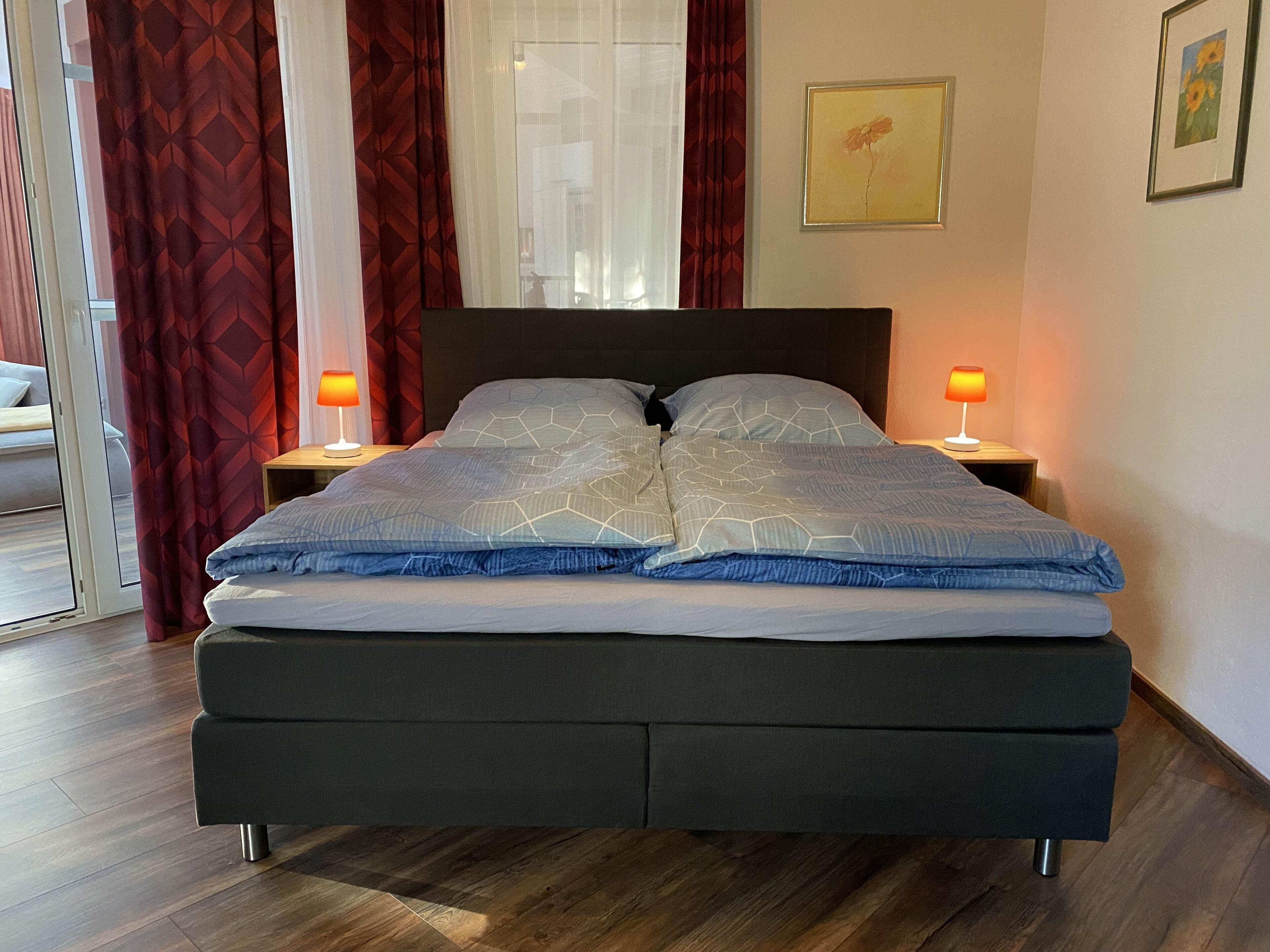 A modern bedroom with a large bed, two bedside tables with lamps, a window with red curtains, and a painting on the wall.