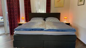 A modern bedroom with a large bed, two bedside tables with lamps, a window with red curtains, and a painting on the wall.