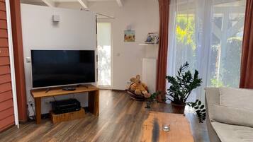 Una sala de estar con una televisión de pantalla plana sobre un mueble de madera, un sofá color beige, una mesa de centro de madera y una planta en maceta.