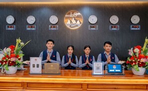 A reception desk with clocks displaying times for various cities, a logo, and staff members. - Lisha roungnakhone hotel (Vang Vieng)