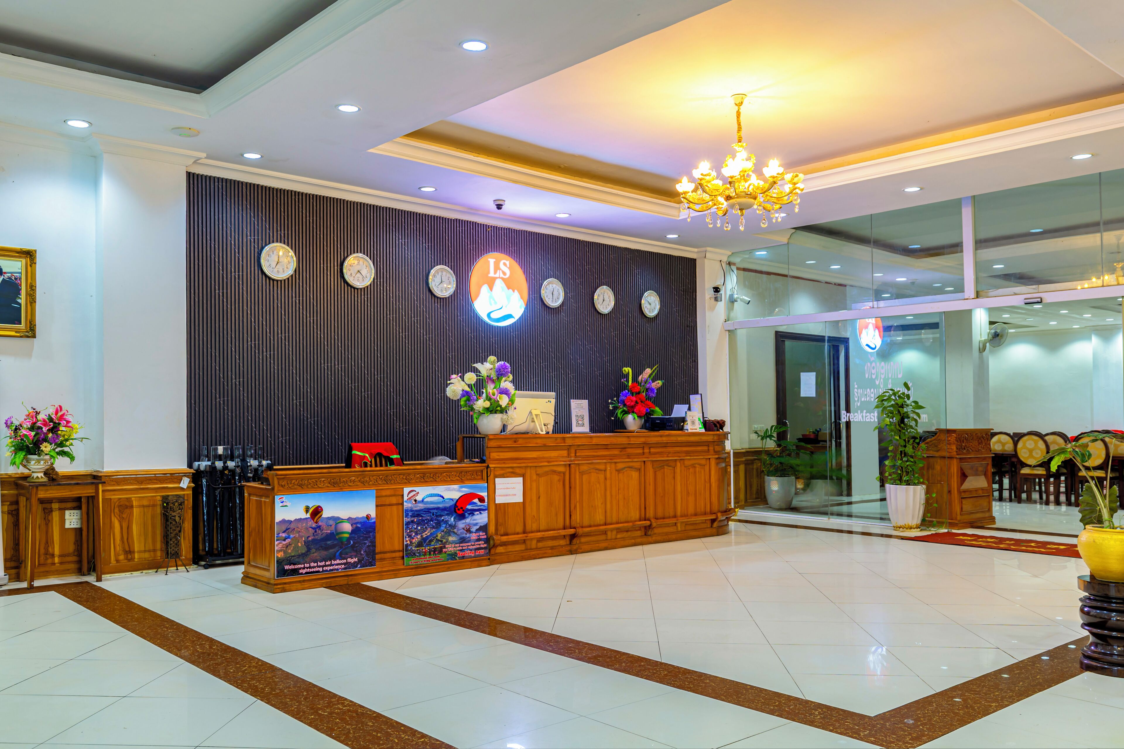 A hotel reception area with a wooden reception desk, multiple clocks, a chandelier, and a large window.