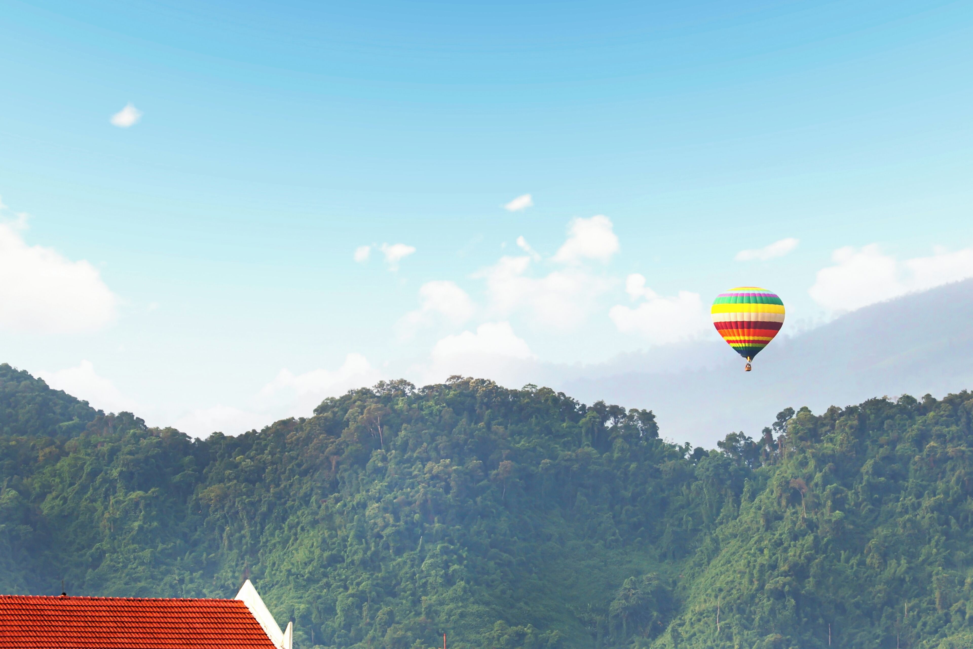 Eine Heißluftballon über einem bewaldeten Hügel, im Vordergrund ein Gebäude mit rotem Dach.