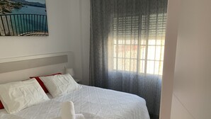 A neatly made bed with white bedding and red accents, a window with sheer curtains, and a painting of a coastal scene above the bed. - Apartment for 2 in Nerja center (Nerja)
