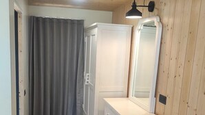 Una habitación pequeña y bien iluminada con paredes y pisos de madera, que incluye un tocador blanco con espejo, un banco y una puerta que da a un baño con cortina.
