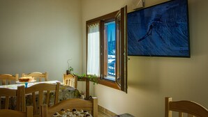 A dining area with a table set for two, a window with a view of a building, and a television mounted on the wall displaying a sports event. - PAESEMIO.CERVAREZZA (Ventasso)
