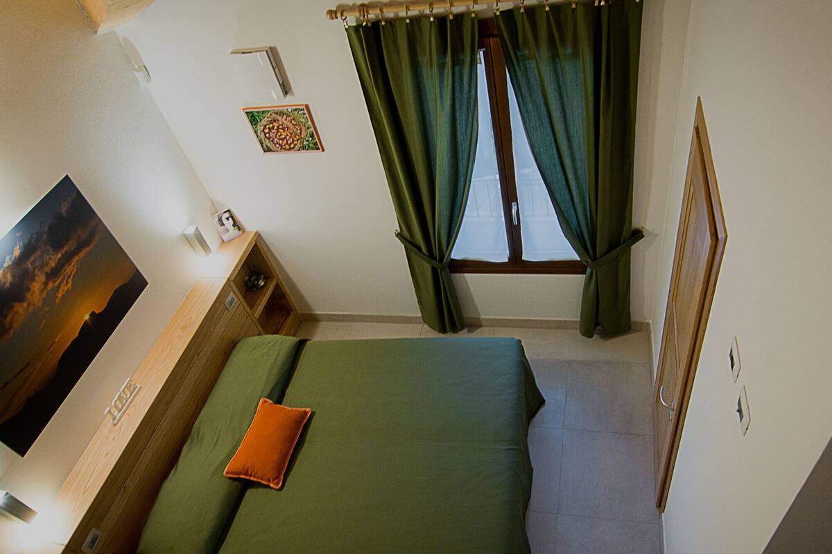 A hotel room with a bed, a nightstand, a wall-mounted TV, a window with curtains, and a framed picture on the wall.