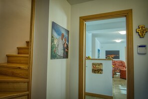 Un pasillo con escaleras de madera, un cuadro, una puerta con un letrero, una cruz y un termostato.