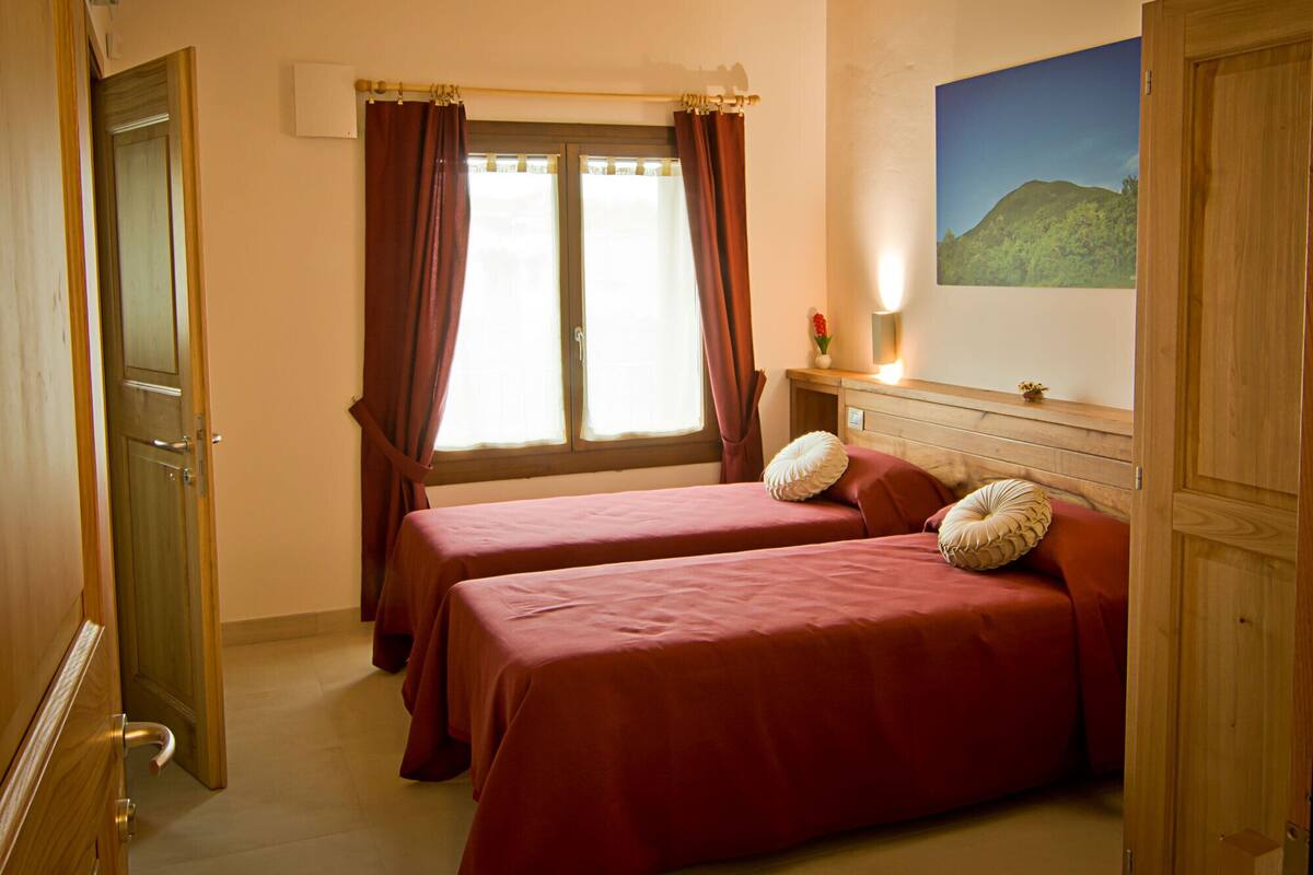 A room with two beds, wooden ceiling beams, a window with curtains, a painting on the wall, and a bedside table with a lamp.