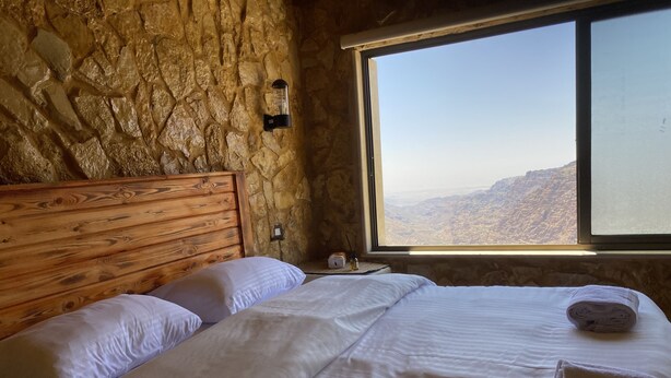Un dormitorio con pared de piedra, cabecero de madera, un ventanal con vistas a la montaña y camas impecablemente preparadas.