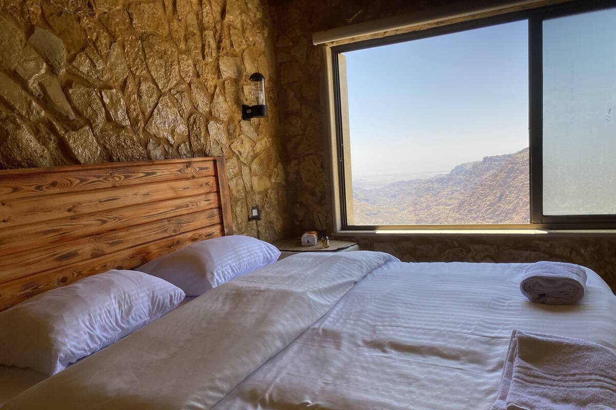 Ein Schlafzimmer mit Steinsturz, Holz-Kopfteil, einem großen Fenster und Blick auf die Berge sowie ordentlich bezogenen Betten.