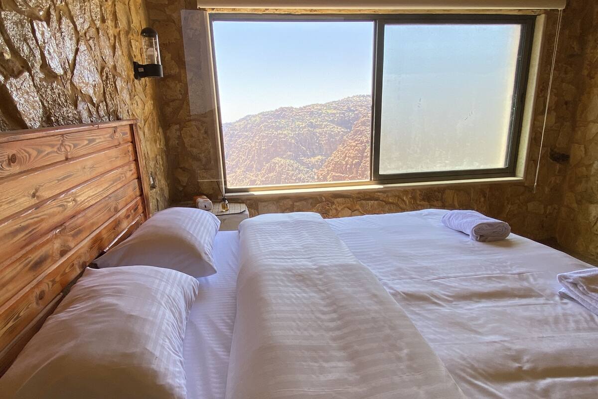 Ein Schlafzimmer mit Holz-Kopfteil, zwei Betten mit weißen Bettwäsche, einem Nachttisch mit Lampe und einem Fenster mit Blick auf die Berge.