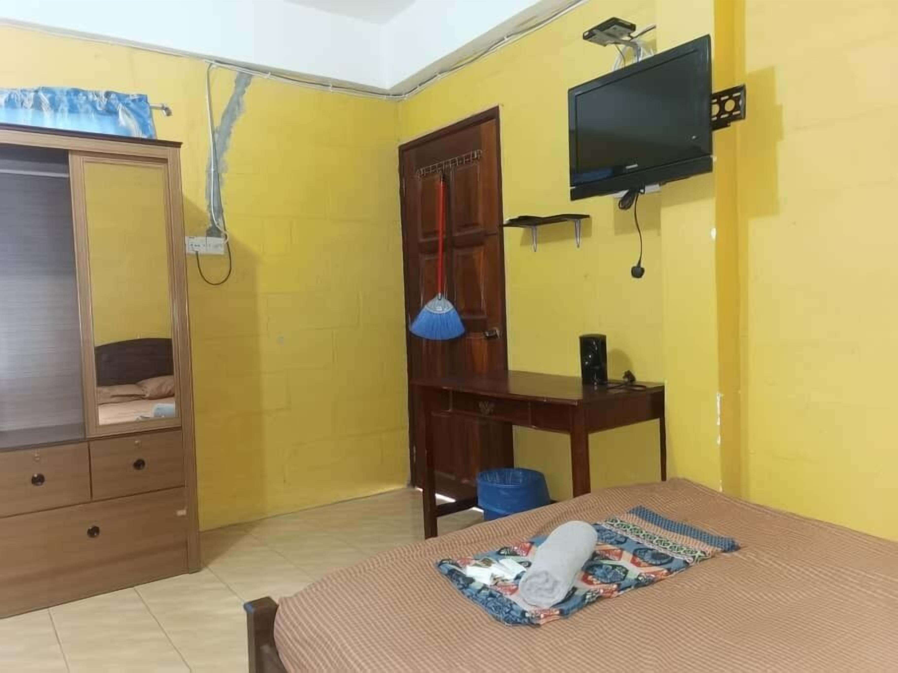 A room with yellow walls, a bed with a patterned bedspread, a wooden wardrobe, a desk with a lamp, and a television mounted on the wall.