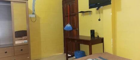 A room with yellow walls, a bed with a patterned bedspread, a wooden wardrobe, a desk with a lamp, and a television mounted on the wall.