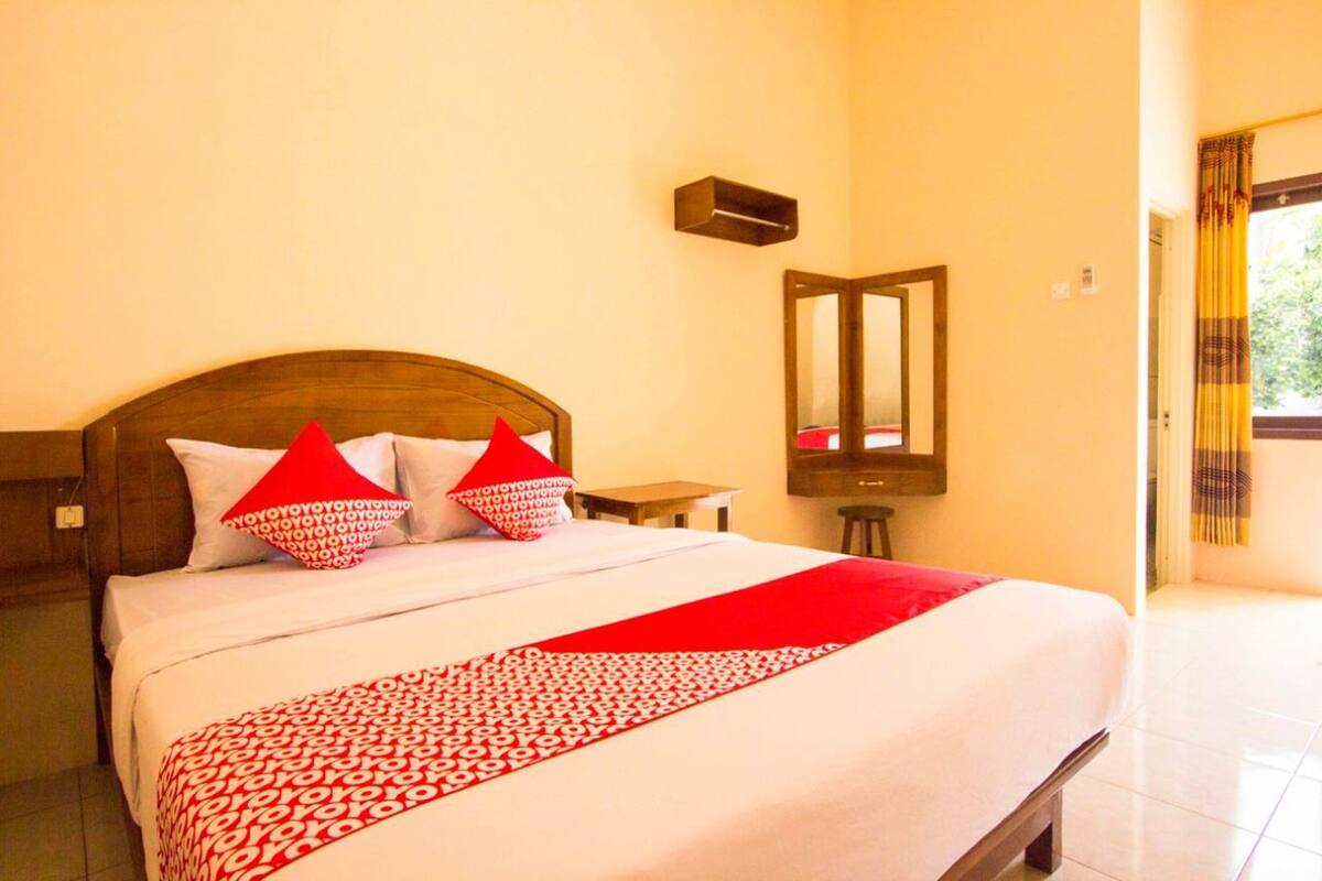 A hotel room with a large bed, a wooden headboard, a small table, a mirror, and a window with curtains.