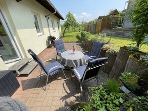 An outdoor patio with a round table and chairs, a barbecue grill, and a stone wall with a wooden gate. - Gemütliche Fewo, Ruhige Lage Direkt Über dem Rheintal Gegenüber der Loreley (Sankt Goar)