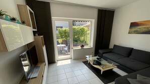 Un salon moderne avec un canapé noir, une table basse en verre et un téléviseur posé sur un meuble en bois. Une porte-fenêtre coulissante donne accès à un espace extérieur aménagé avec une table et des chaises.