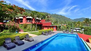 A pool area with lounge chairs, a red building with multiple levels, and a mountainous backdrop. - Villa Noelia Garden Suite - Coatepeque Lake (Lago de Coatepeque)