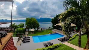 Un espace piscine avec des chaises longues, un toboggan aquatique en forme de cœur et une vue sur un lac bordé de montagnes.
