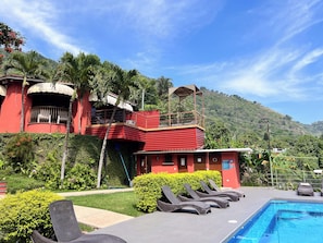 A red building with a balcony, a swimming pool with lounge chairs, and a lush green landscape.