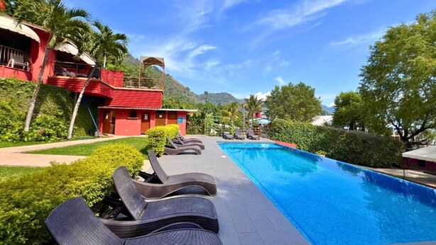 A swimming pool with lounge chairs, a red building with a balcony, and lush greenery. - Villa Noelia Calli Suite - Coatepeque Lake (Lago de Coatepeque)
