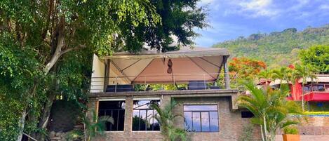 Ein zweistöckiges Gebäude mit einer überdachten Terrasse, einer Garage und Blick auf eine hügelige Landschaft.