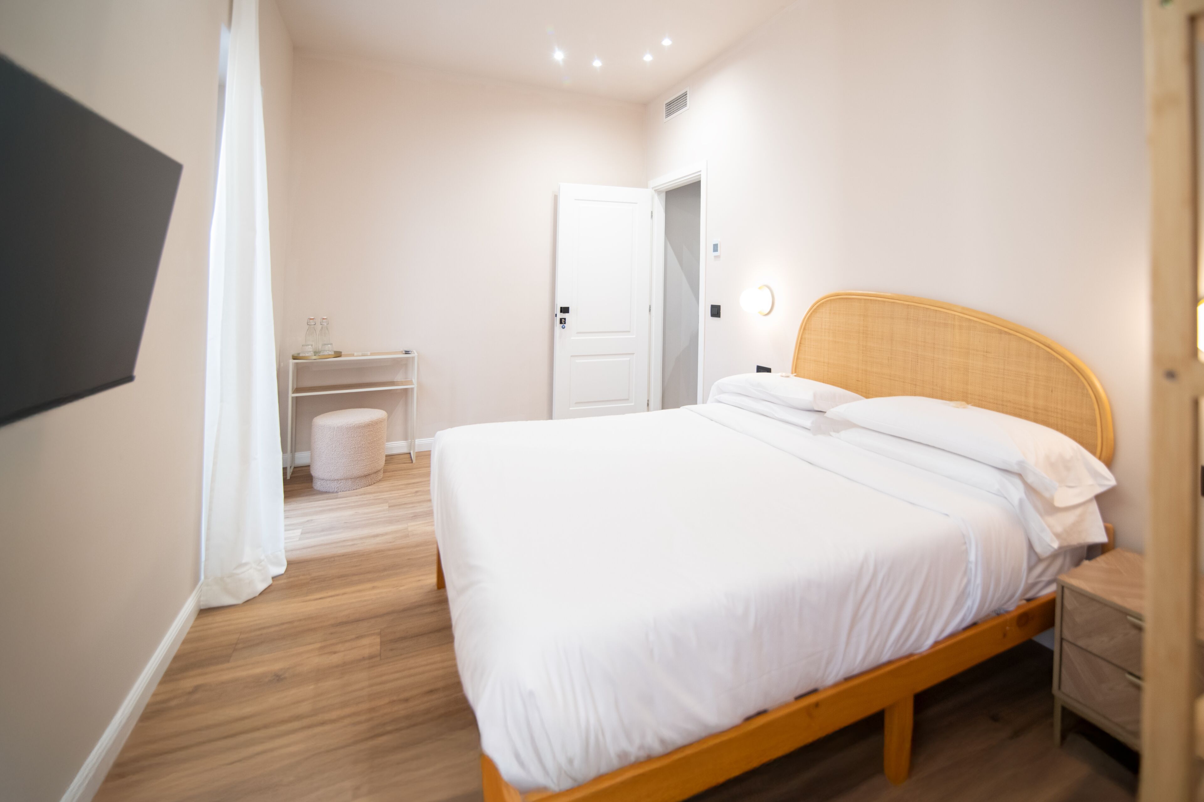 A neatly arranged bedroom with a wooden bed, a bedside table, a small shelf with bottles, and a white door.
