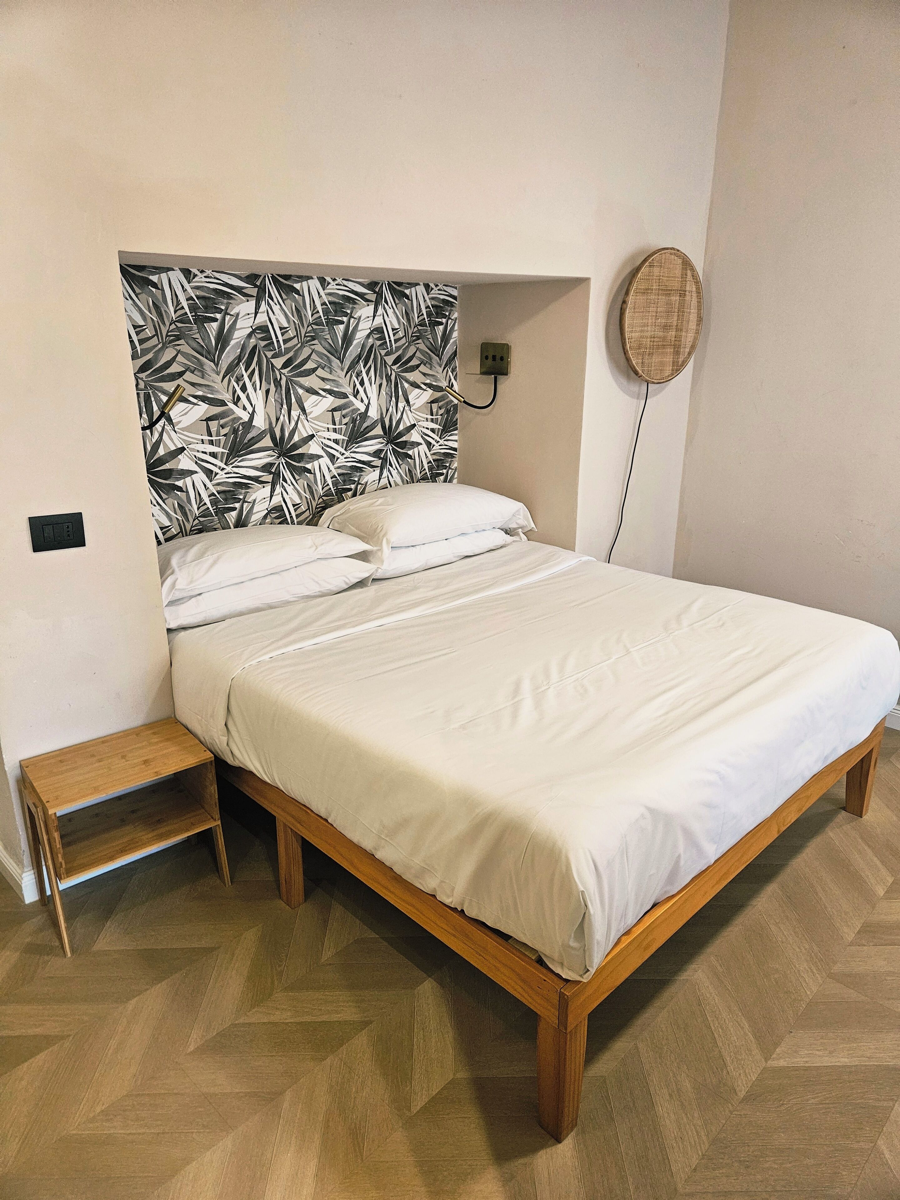 Un lit avec un cadre en bois et une literie blanche, une petite table de chevet en bois, une table ronde fixée au mur et une tête de lit recouverte d’un papier peint à motifs.