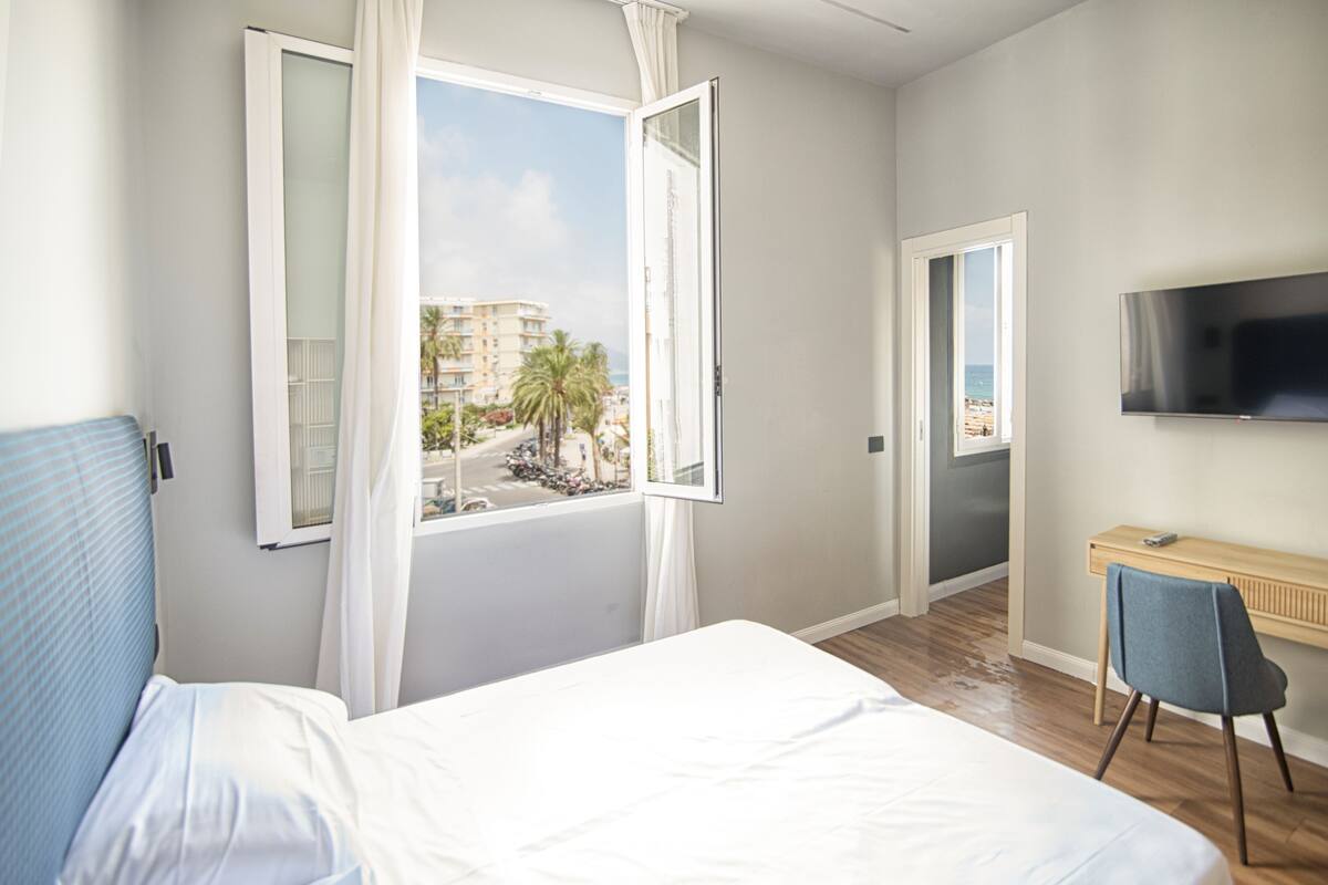 Une chambre d’hôtel équipée d’un lit, d’un bureau, d’une chaise, d’une télévision et d’une fenêtre donnant sur des immeubles et des palmiers.