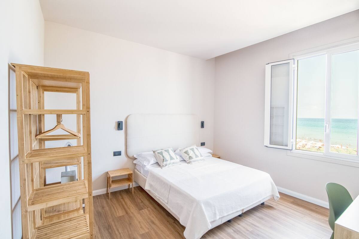 Une chambre à coucher comprenant un lit, une étagère en bois, une table de chevet, une chaise et une fenêtre donnant sur la plage.