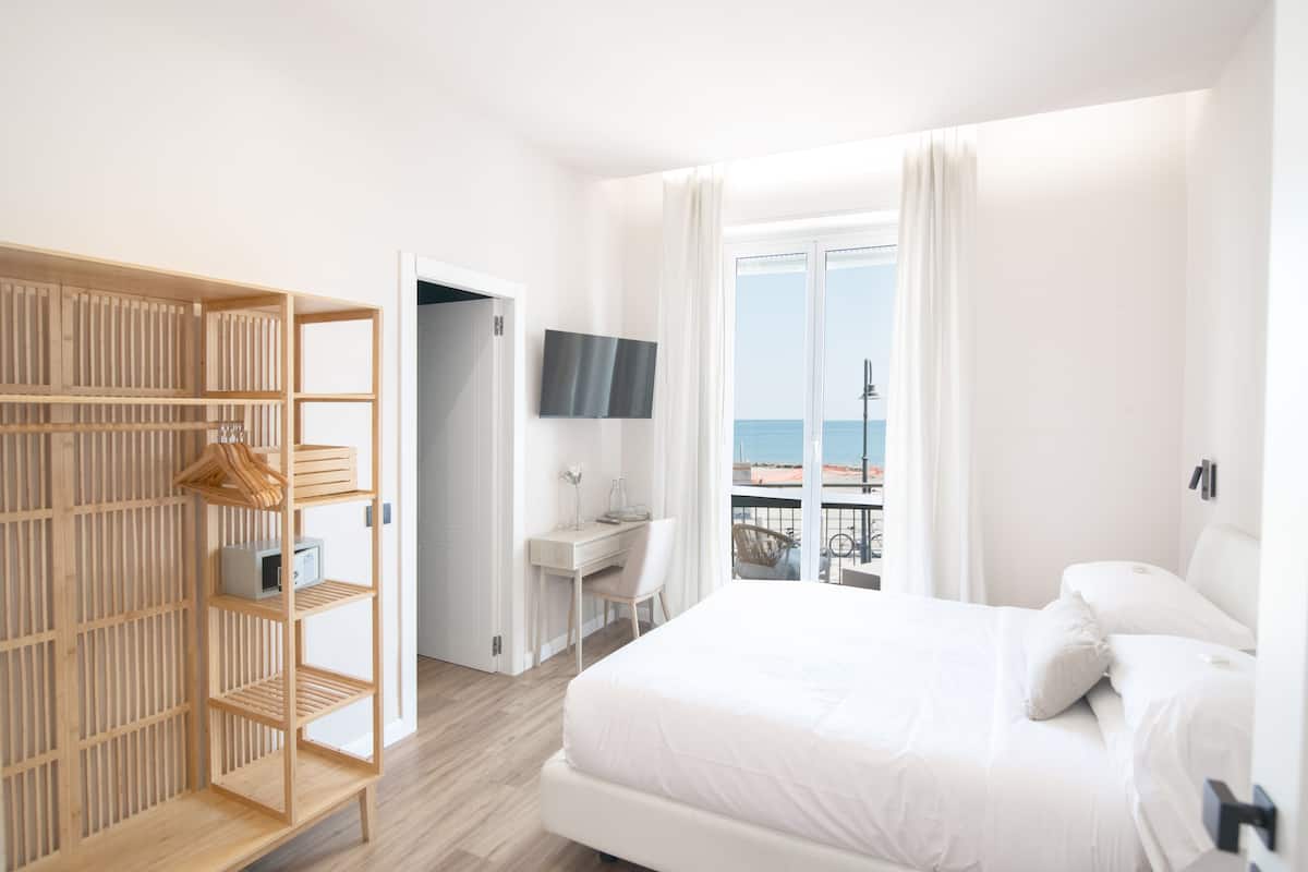 Une chambre d’hôtel moderne dotée d’un grand lit, d’une armoire en bois, d’un bureau avec une chaise, d’une télévision et d’un balcon offrant une vue sur la mer.