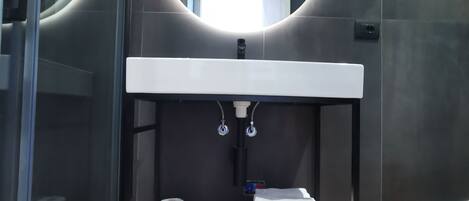Un baño moderno con un espejo redondo, un lavamanos blanco y un estante con toallas y papel higiénico.