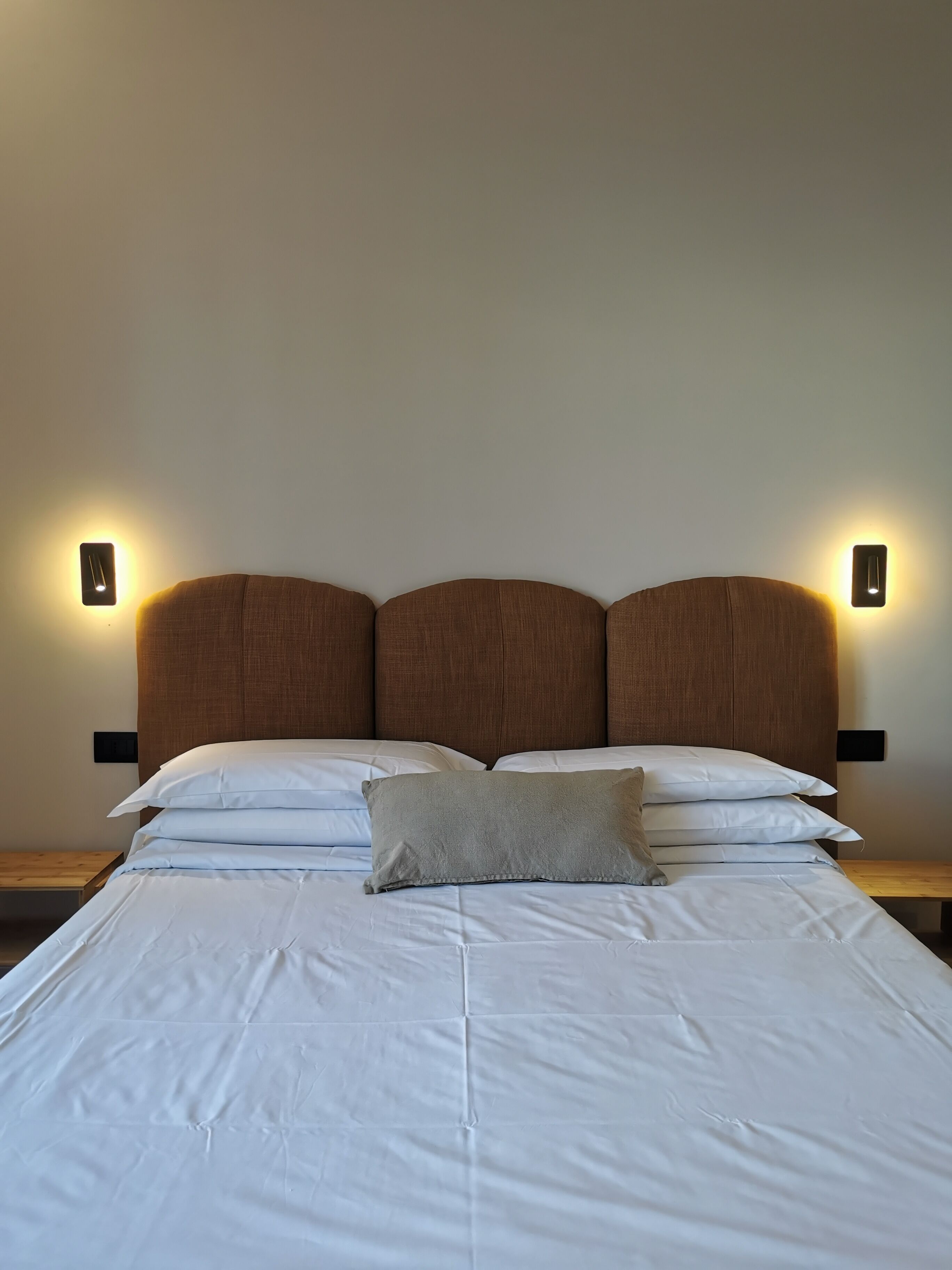 Una cama bien hecha con sábanas blancas y una sola almohada, flanqueada por dos lámparas fijadas a la pared.