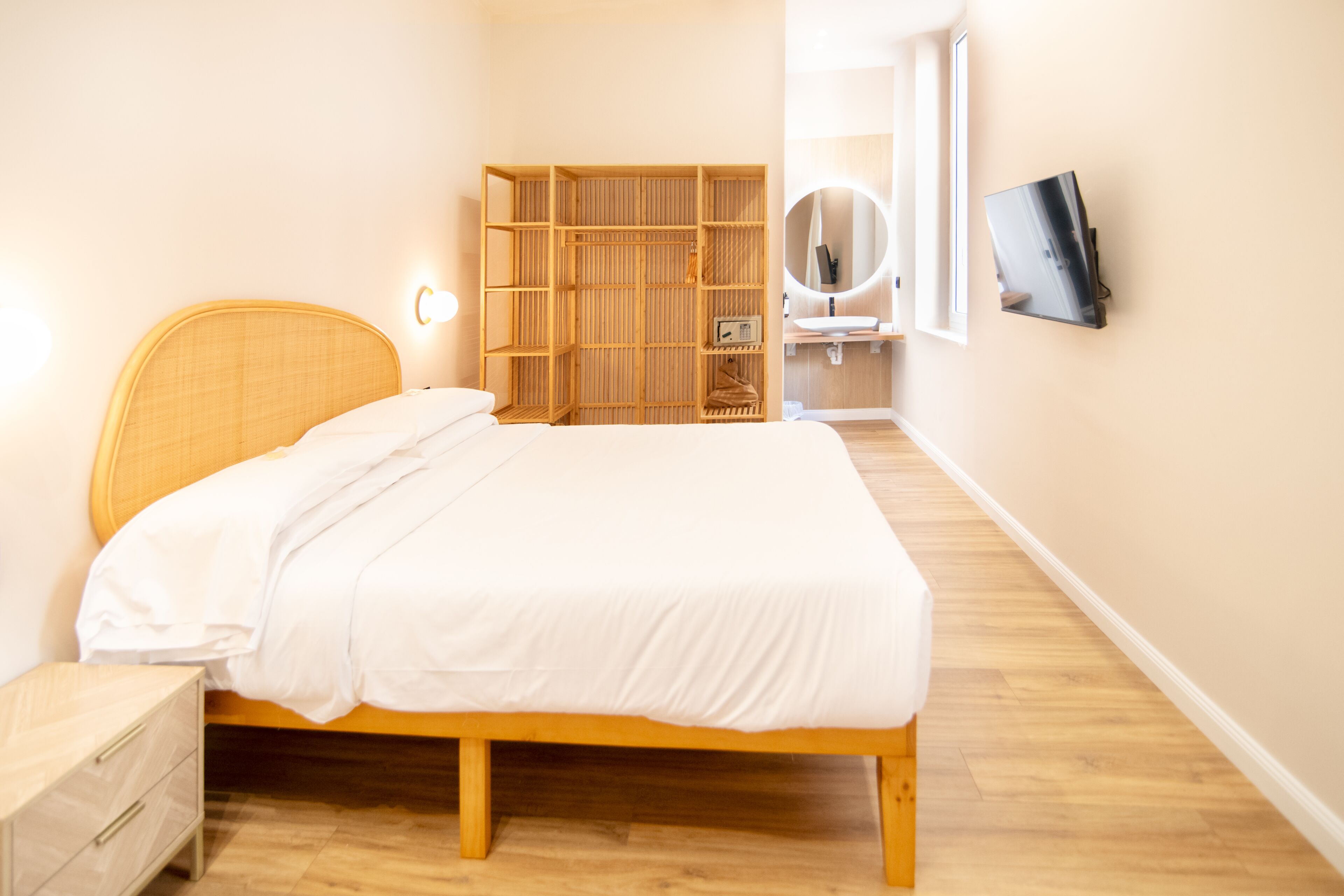 Une chambre à coucher avec un lit en bois, une armoire, une table de chevet, un téléviseur fixé au mur et un miroir au-dessus du lavabo.