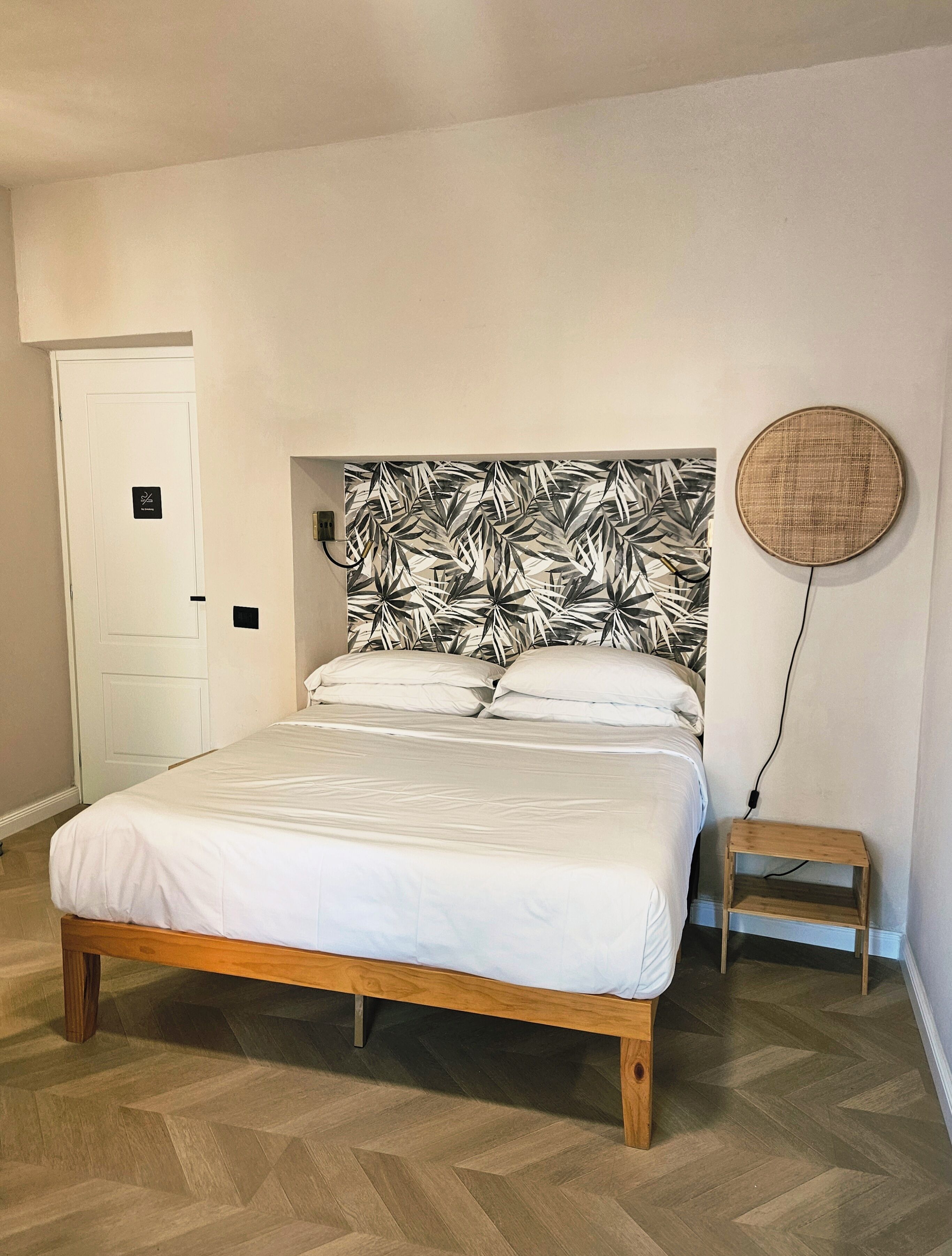 Une chambre à coucher avec un lit, une petite table de chevet en bois, une lampe ronde fixée au mur et une porte avec une pancarte.
