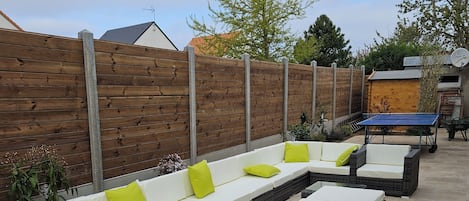 A patio area with a long white sofa with green cushions, a ping pong table, and a small garden area with plants.