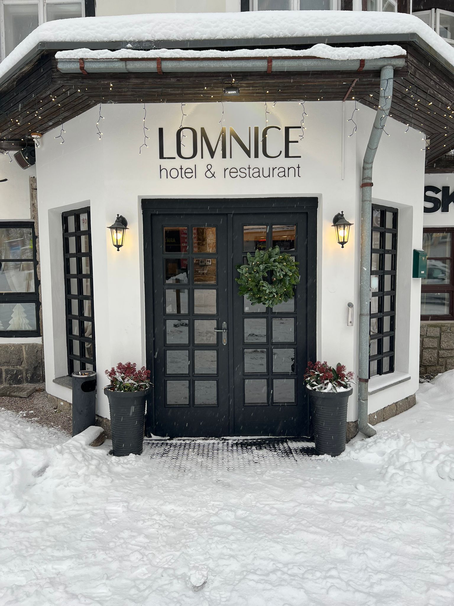 A white building with a black door, labeled "LOMNICE hotel & restaurant," featuring a wreath and potted plants.