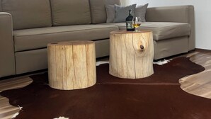 Una sala moderna con un sofá gris, dos taburetes de madera, un tapete de piel de vaca y una botella de licor sobre una mesa redonda de madera.