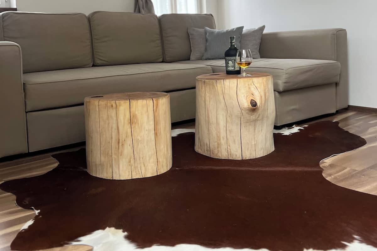 A modern living room with a grey sofa, two wooden stools, a cowhide rug, and a bottle of liquor on a round wooden table.