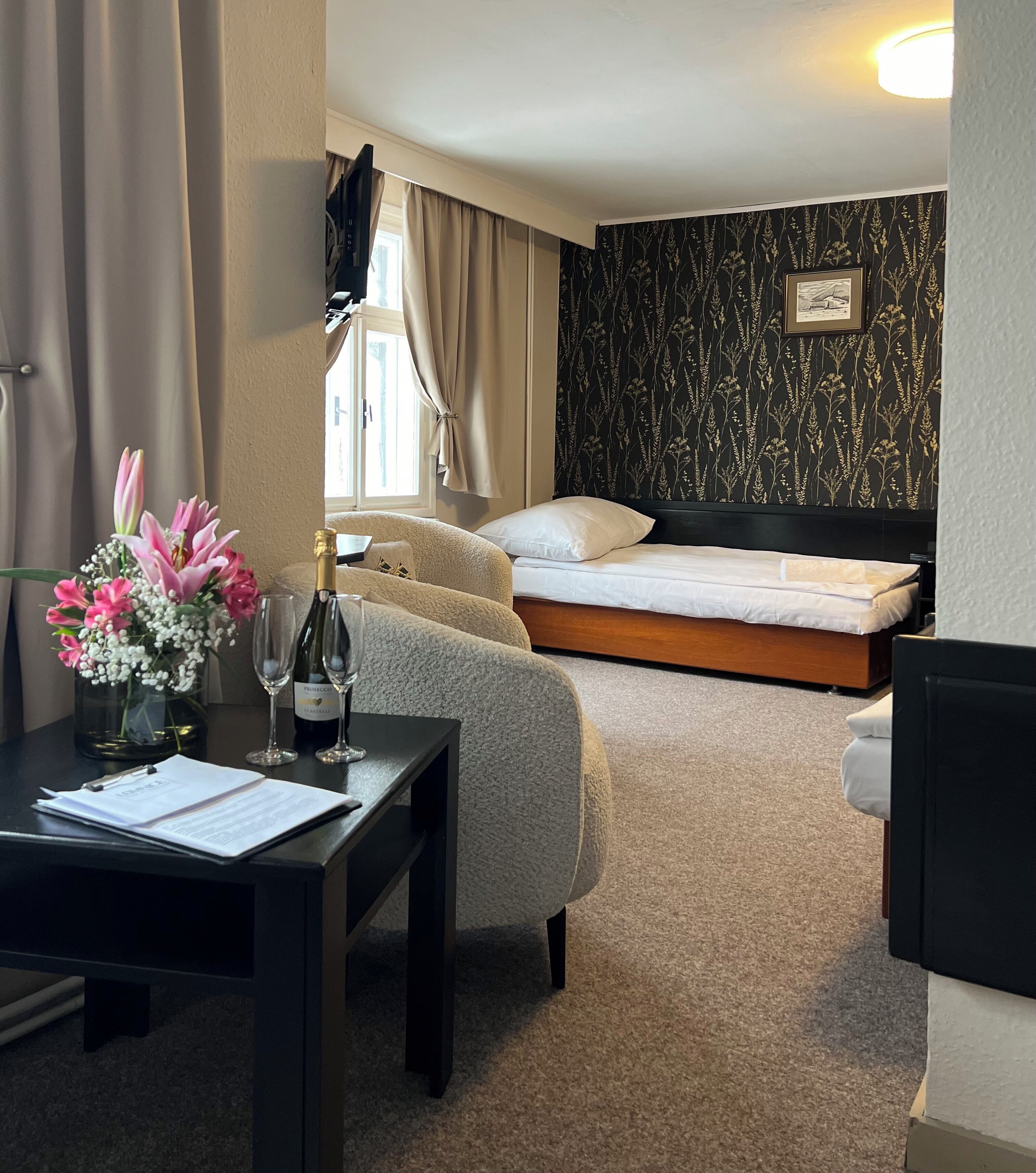 Una habitación de hotel con una cama, un escritorio con un florero, una botella de champán y un cuadro en la pared.