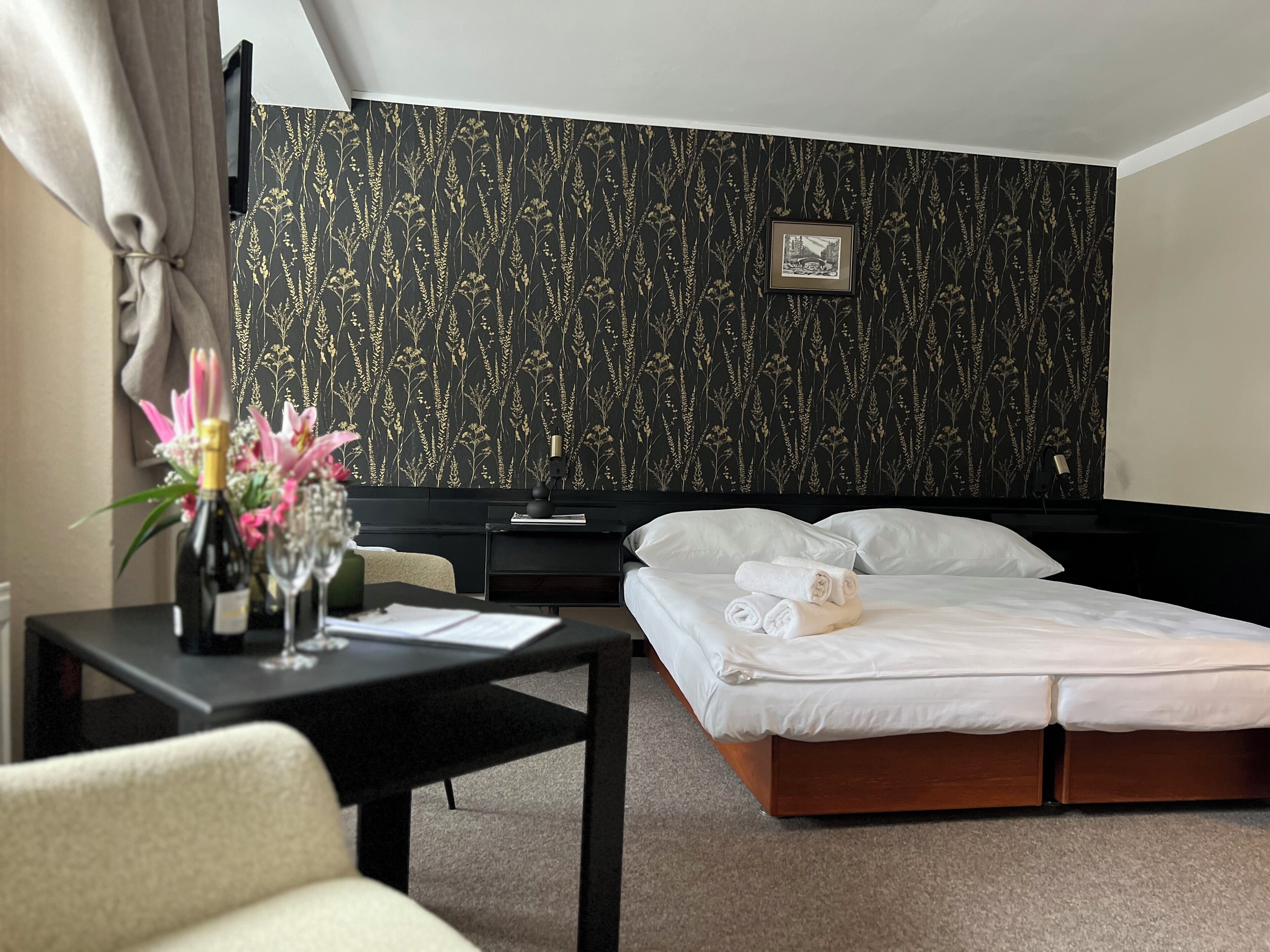 A hotel room with a bed, a desk with a lamp, a chair, and a small table with a vase of flowers.