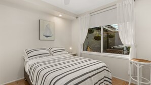 Un dormitorio con cama, mesita de noche, ventana con cortinas y una planta colgante.