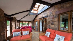 A spacious indoor area with stone walls and a high ceiling. There are two wicker sofas with cushions, a wooden table, and a glass door leading outside. - Lakeside Escape, Stone Countryside Cottage (Leek)