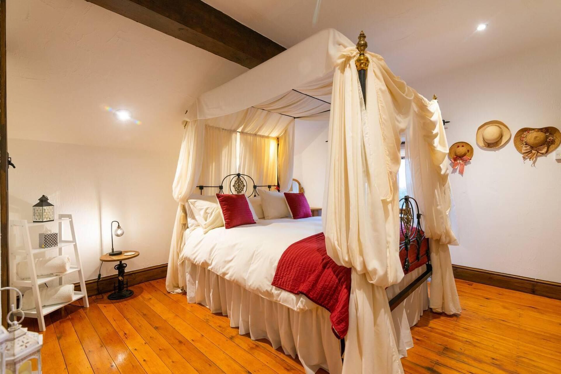 A four-poster canopy bed with white and red bedding, a wooden floor, and a white wall with a small shelf and two hats.