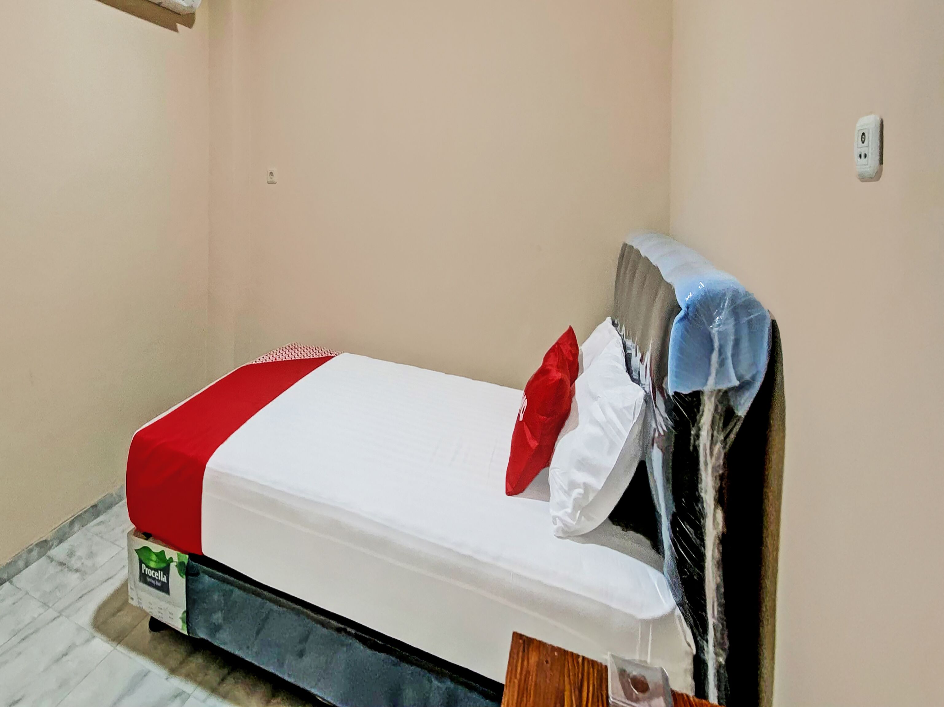 Ein kleines Zimmer mit einem Einzelbett, einer weißen Matratze, rot-weißer Bettwäsche und einem Matratzenschoner.